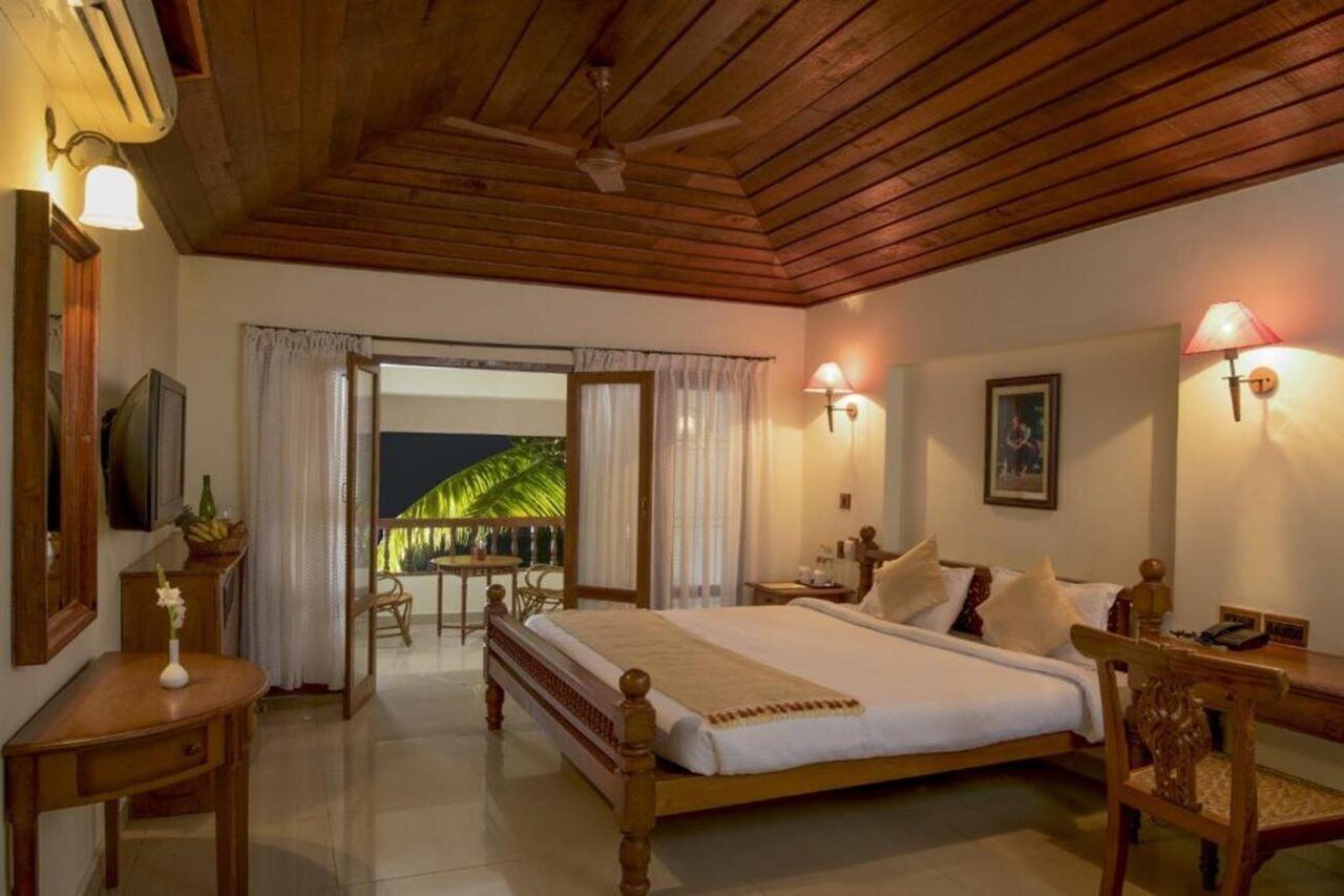 A bedroom with a wooden ceiling, a bed with white linens, a desk with a lamp, and a view of a palm tree through the open door.