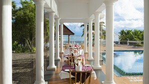 An outdoor dining area with white columns, tables set with purple tablecloths, and a view of the ocean. - Isola di Cocco Ayurvedic Beach Resort (Neyyattinkara)