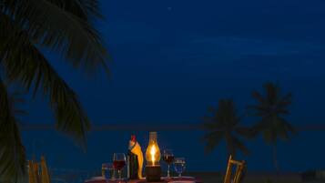 A table set for dining with glasses and a bottle, palm trees in the background, and a night sky.