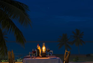 A table set for dining with glasses and a bottle, palm trees in the background, and a night sky.