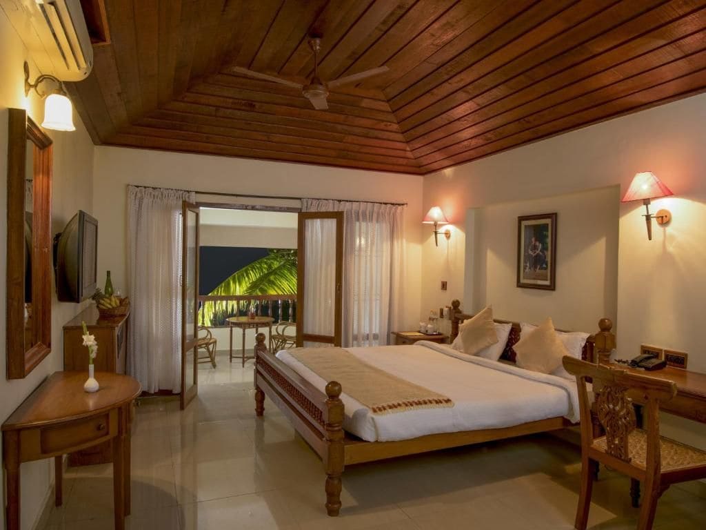A bedroom with a wooden ceiling, a bed with white bedding, a desk with a lamp, and a view of greenery outside.