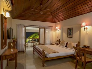 A bedroom with a wooden ceiling, a bed with white bedding, a desk with a lamp, and a view of greenery outside. - Isola di Cocco Ayurvedic Beach Resort (Neyyattinkara)