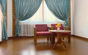 Ein Zimmer mit einer Couch, zwei Stühlen, einem kleinen Tisch mit Blumenvase und einem fenster mit Vorhängen.