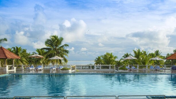 A swimming pool with lounge chairs and umbrellas, surrounded by palm trees and a view of the ocean. - Isola di Cocco Ayurvedic Beach Resort (Neyyattinkara)