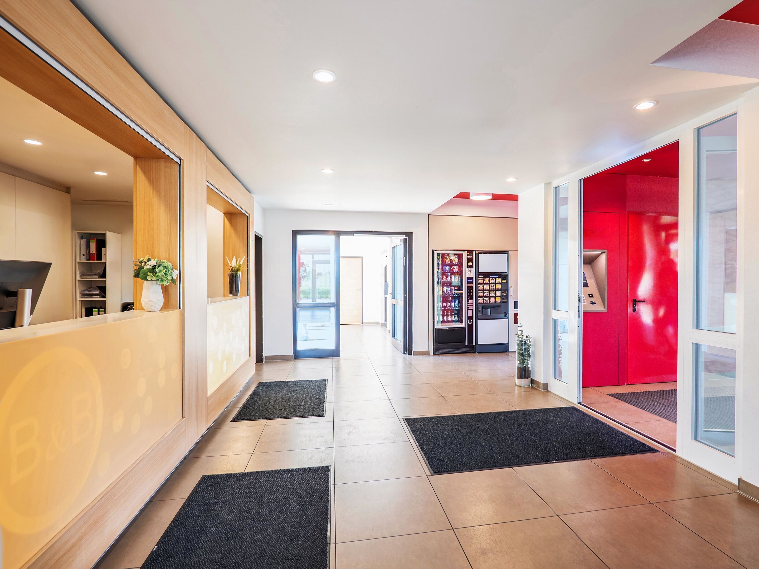 A modern hotel lobby with a reception desk, a vending machine, and a red accent wall.