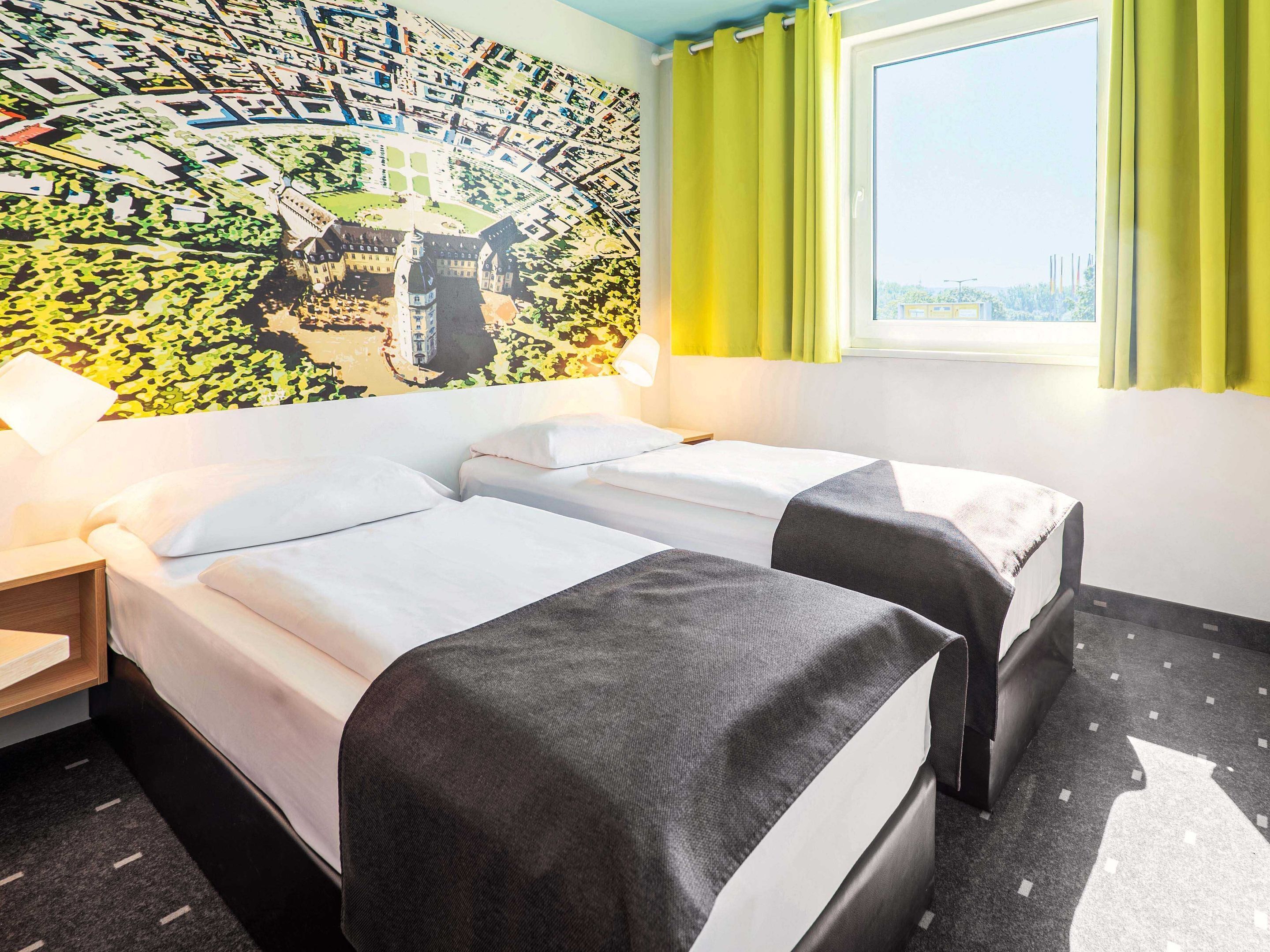 Een hotelkamer met twee bedden, een groot raam met gordijnen, een stadslandschap muurschildering op de muur, en een nachtkastje met een lamp.
