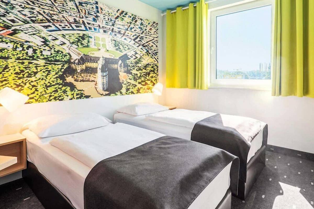 Ein Hotelzimmer mit zwei Betten, einem Stadtbild-Wandgemälde, einem Fenster mit Vorhängen und einem Nachttisch.