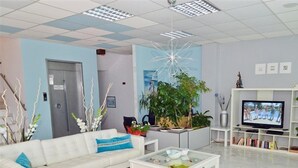 A modern living room with a white sofa, a glass coffee table, and a TV on a white stand. There are plants and a painting on the wall. - Astris Sun (Thasos)