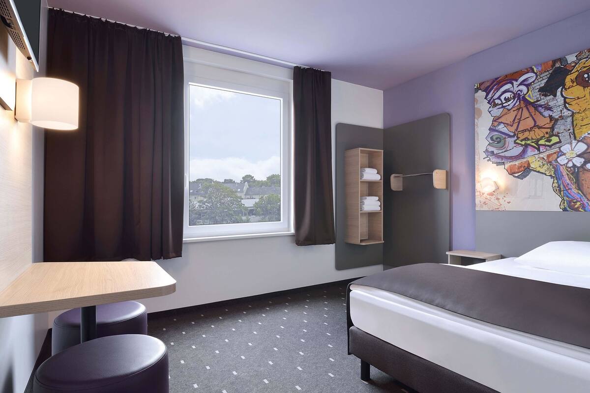Moderni hotellihuone, jossa on sänky, työpöytä, tuoli, ikkuna verhoilla, seinälle kiinnitetty hylly pyyhkeille ja värikäs abstrakti maalaus.