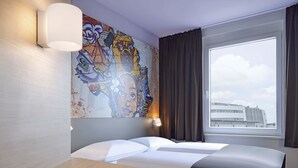 A hotel room with a bed, a desk, a chair, a large window with curtains, and a colorful mural on the wall. - B&B HOTEL Köln-Ehrenfeld (Cologne)