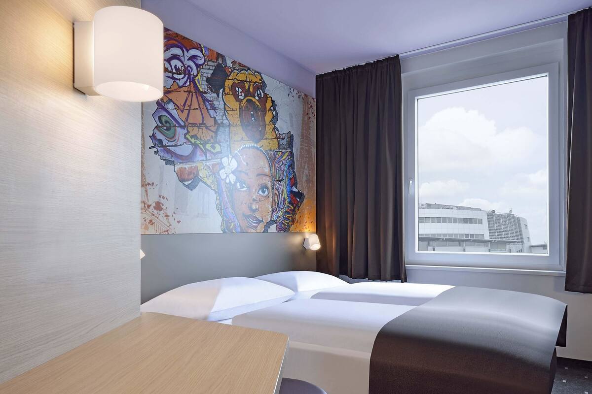 Hotellihuone, jossa on sänky, työpöytä, tuoli, suuri ikkuna verhoilla sekä värikäs seinämaalaus.