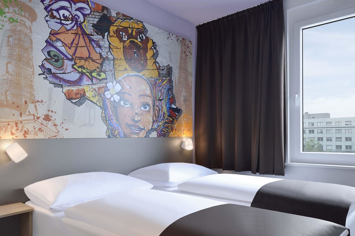 Hotellihuone, jossa on sänky, yöpöytä, suuri seinämaalaus, ikkuna verhoilla ja näkymä ulkona oleviin rakennuksiin.