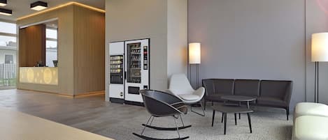 A modern hotel lobby with a reception desk, vending machine, seating area, and a large wooden table.