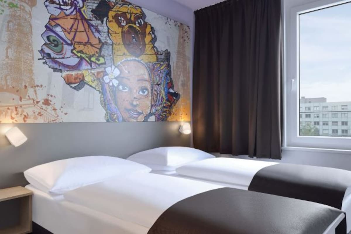 A hotel room with a bed, a nightstand, a wall mural, and a window with curtains.