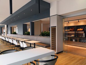Un espace de restauration moderne avec de longues tables et des chaises, un comptoir où sont exposés les plats, et un coin salon agrémenté de plantes.