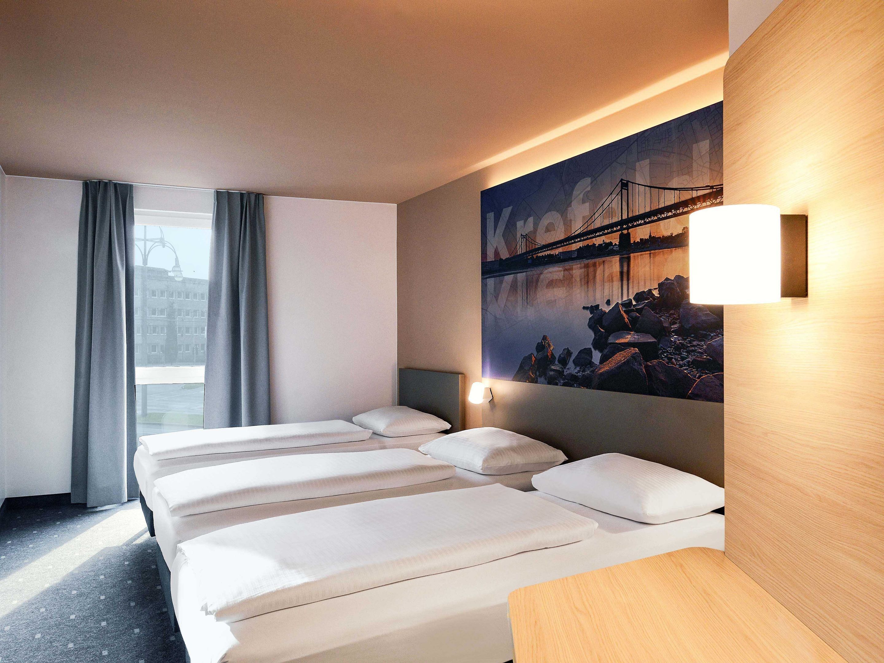 Habitación de hotel con cuatro camas, un cuadro grande de un puente, una ventana con cortinas y una lámpara fijada a la pared.