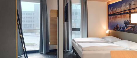 Una habitación de hotel con cama, escritorio, televisión y un amplio ventanal con cortinas.