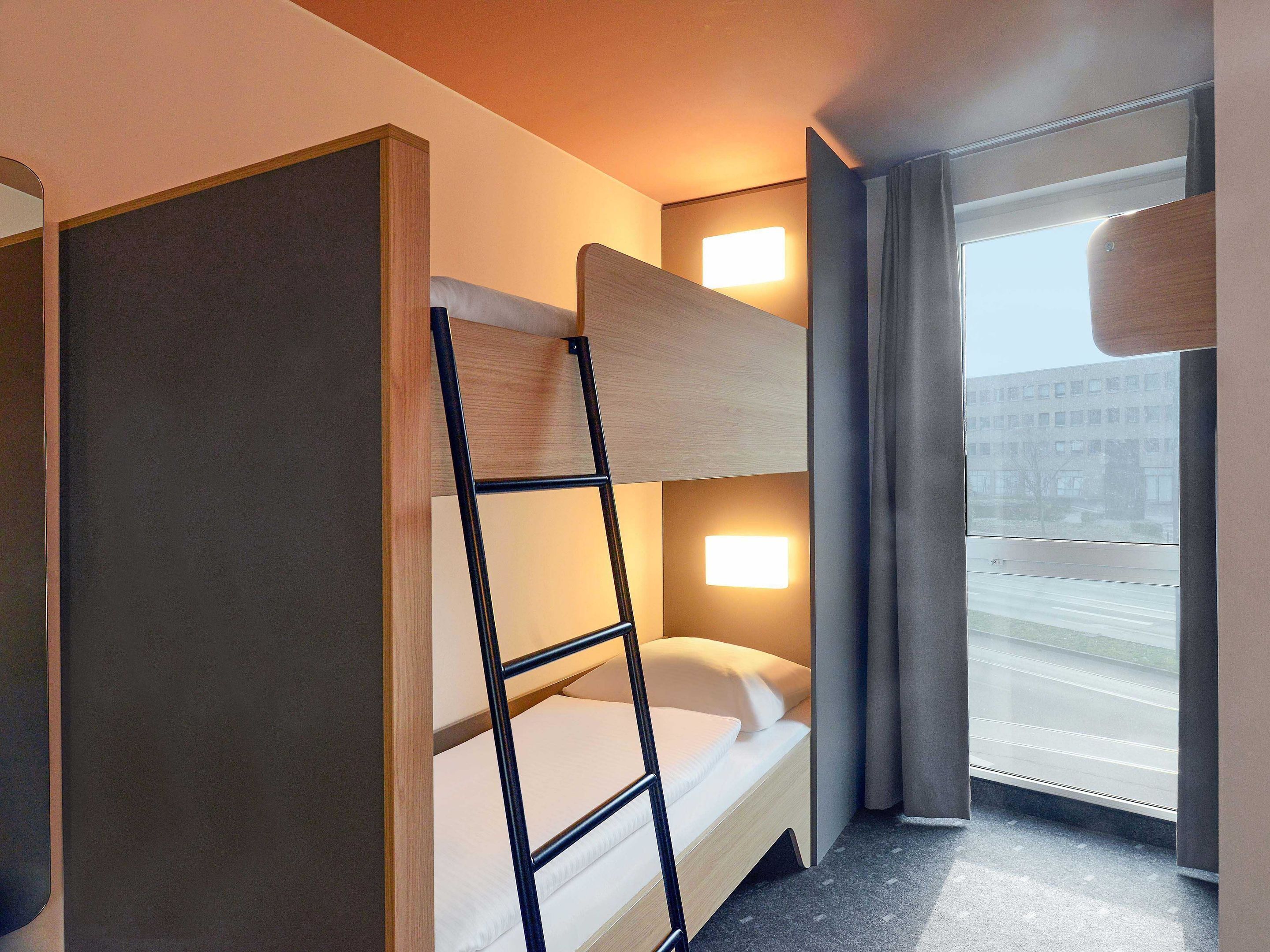 Een kleine hotelkamer met een stapelbed, nachtkastje, lamp en een raam met gordijnen.