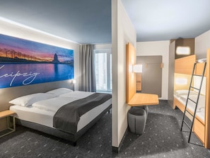 Habitación de hotel con una cama grande, una mesita pequeña, una mesita de noche, una lámpara de pared, una ventana con cortinas y vistas a un paisaje urbano.