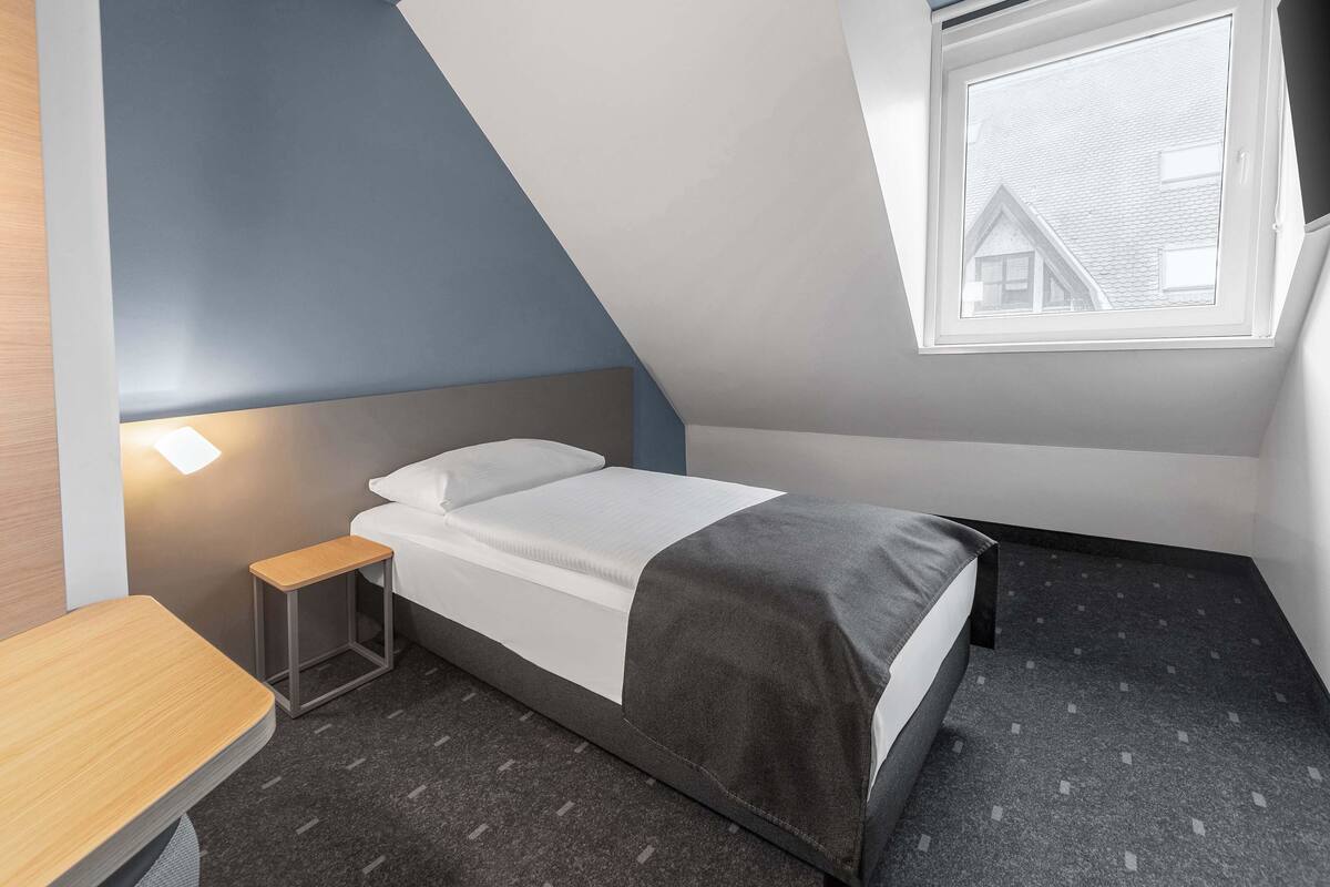 A small, modern bedroom with a bed, a bedside table, and a window.