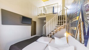 Ein modernes Hotelzimmer mit Bett, Fernseher, einer Treppe und einem Stadtbild-Wandgemälde.