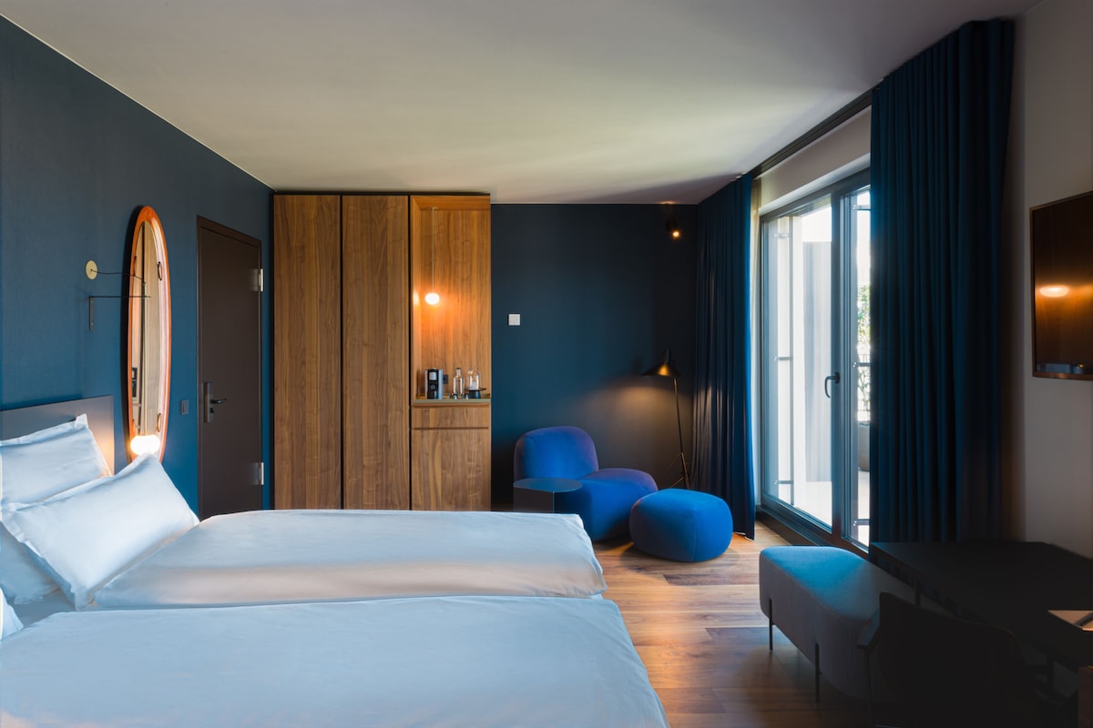 Una habitación de hotel moderna con una cama grande, un armario de madera, una zona de estar con sillas azules y un ventanal con cortinas.