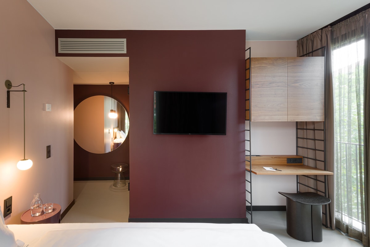 Habitación de hotel moderna con televisión de pantalla plana, espejo redondo, mesita de noche, lámpara y vistas al exterior.