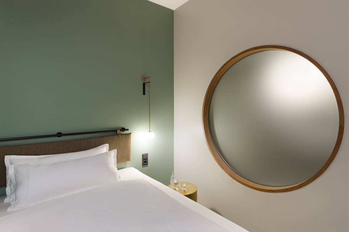 A hotel room with a bed, a round mirror, a bedside table with a lamp, and a wall-mounted light.
