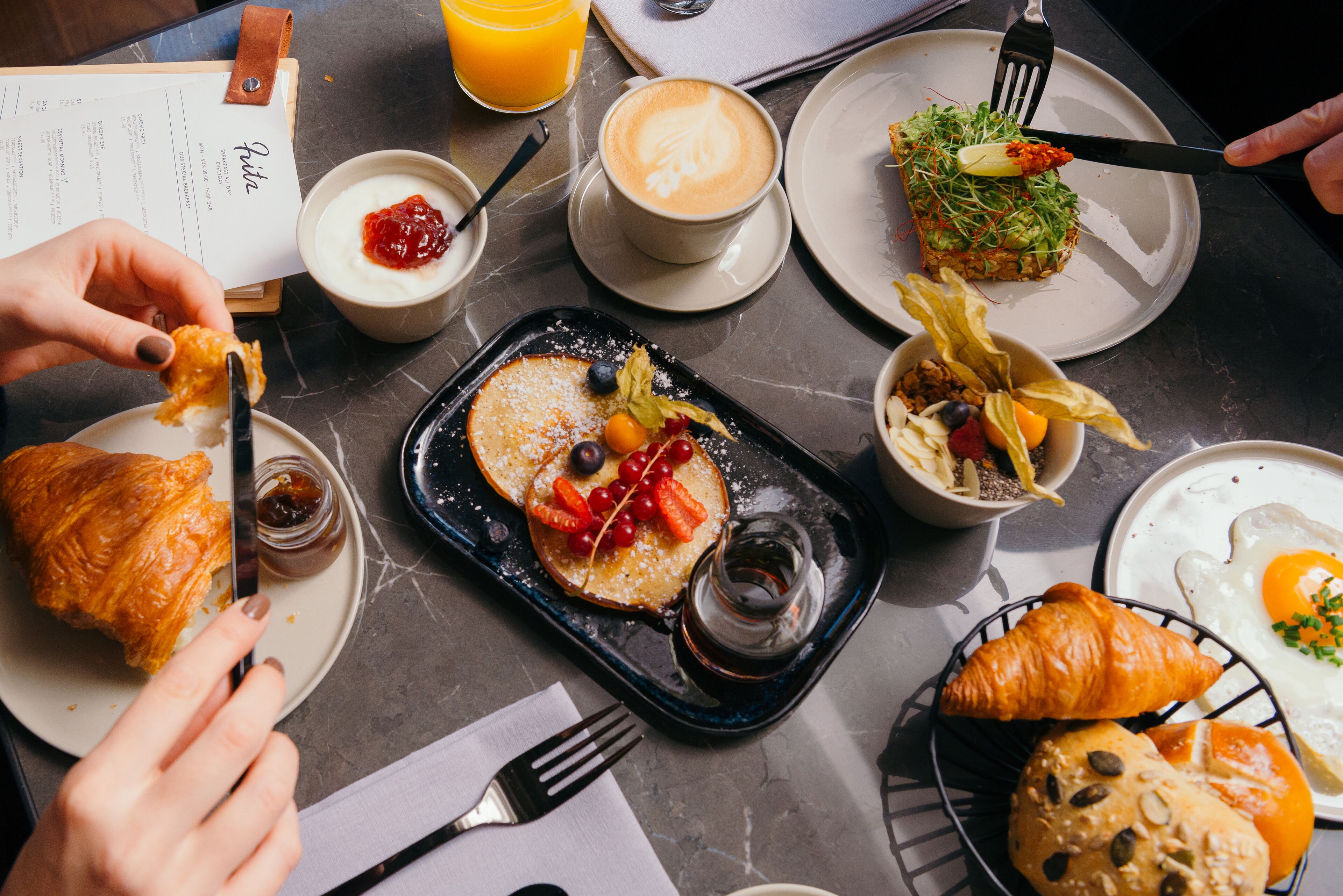 A breakfast setting with croissants, pancakes, coffee, and various fruits on a marble table.