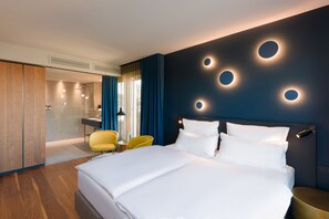 A modern hotel room with a large bed, a bedside table, a chair, and a bathroom visible through a glass partition. - EmiLu Design Hotel (Stuttgart)