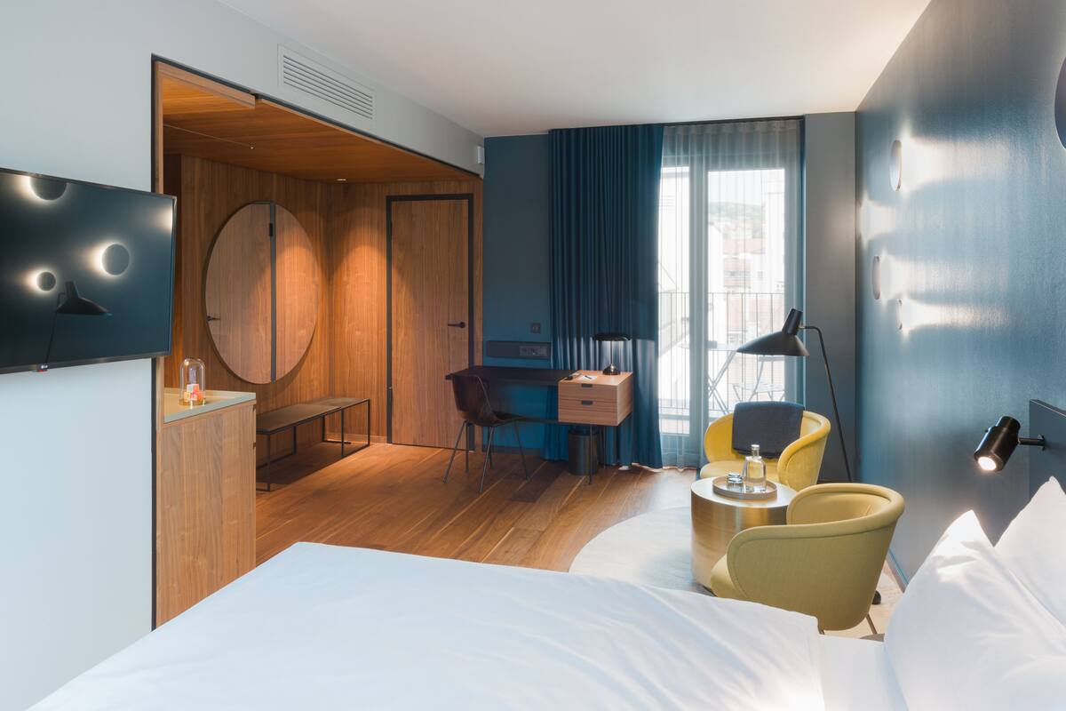 A modern hotel room with a bed, a desk, a chair, a small table, and a TV.