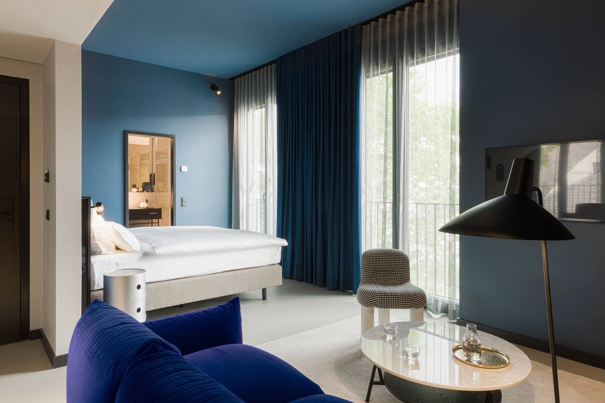 A modern hotel room with a bed, a blue sofa, a round coffee table, a chair, and a floor lamp.