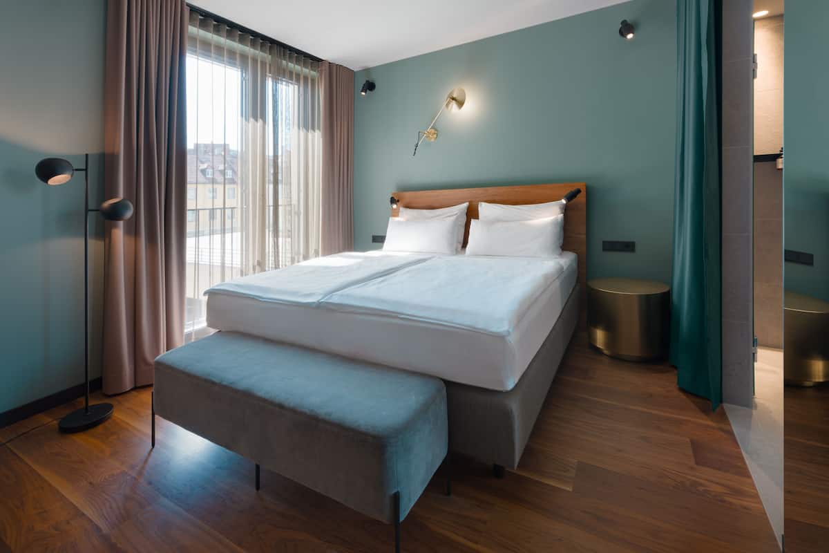 A modern hotel room with a large bed, a bench, a floor lamp, and a view of the city through the window.