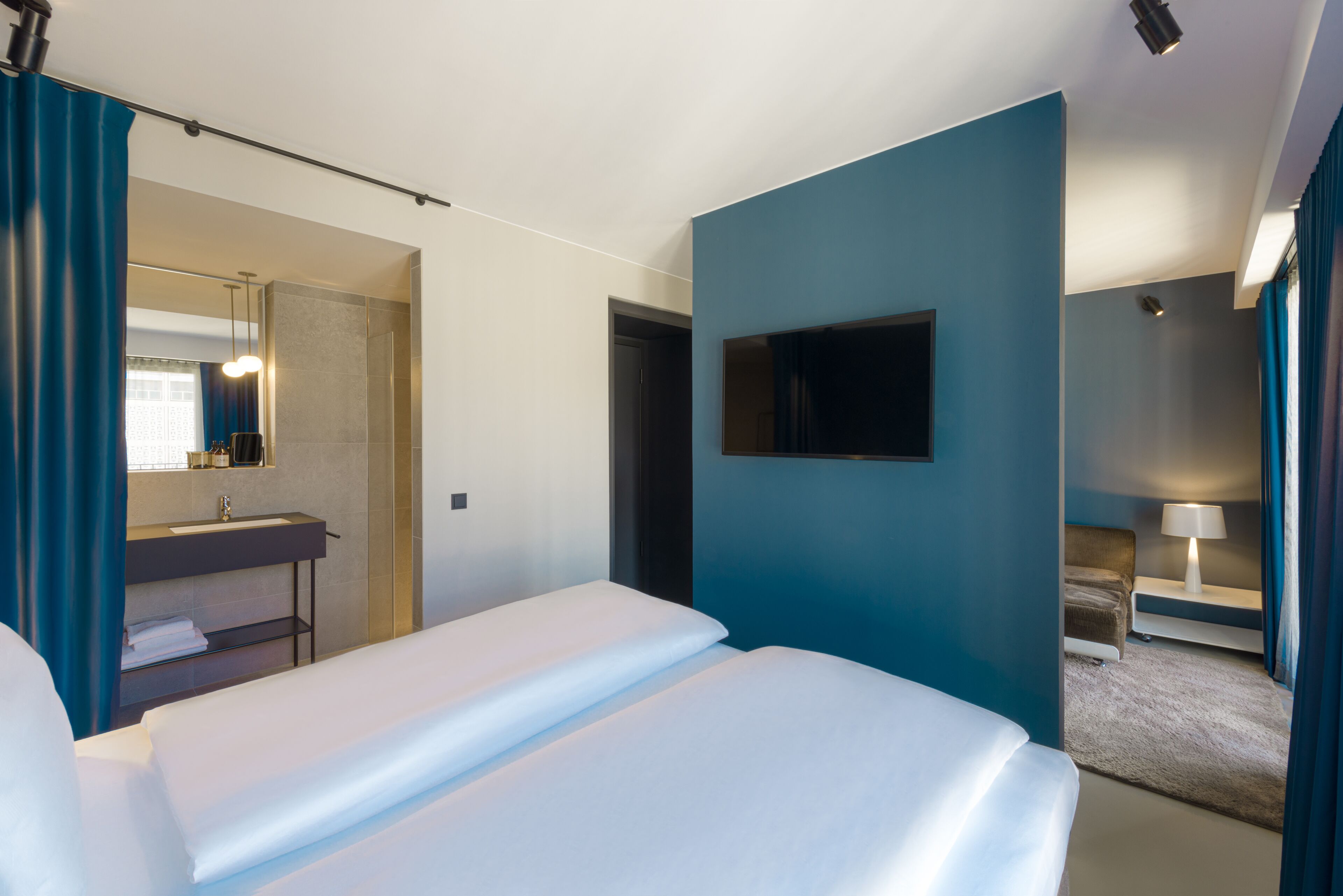 Habitación de hotel con cama, televisor en la pared, baño con lavamanos y espejo, y una pequeña zona de estar con una lámpara.