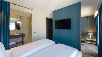 Habitación de hotel con cama, televisor en la pared, baño con lavamanos y espejo, y una pequeña zona de estar con una lámpara.