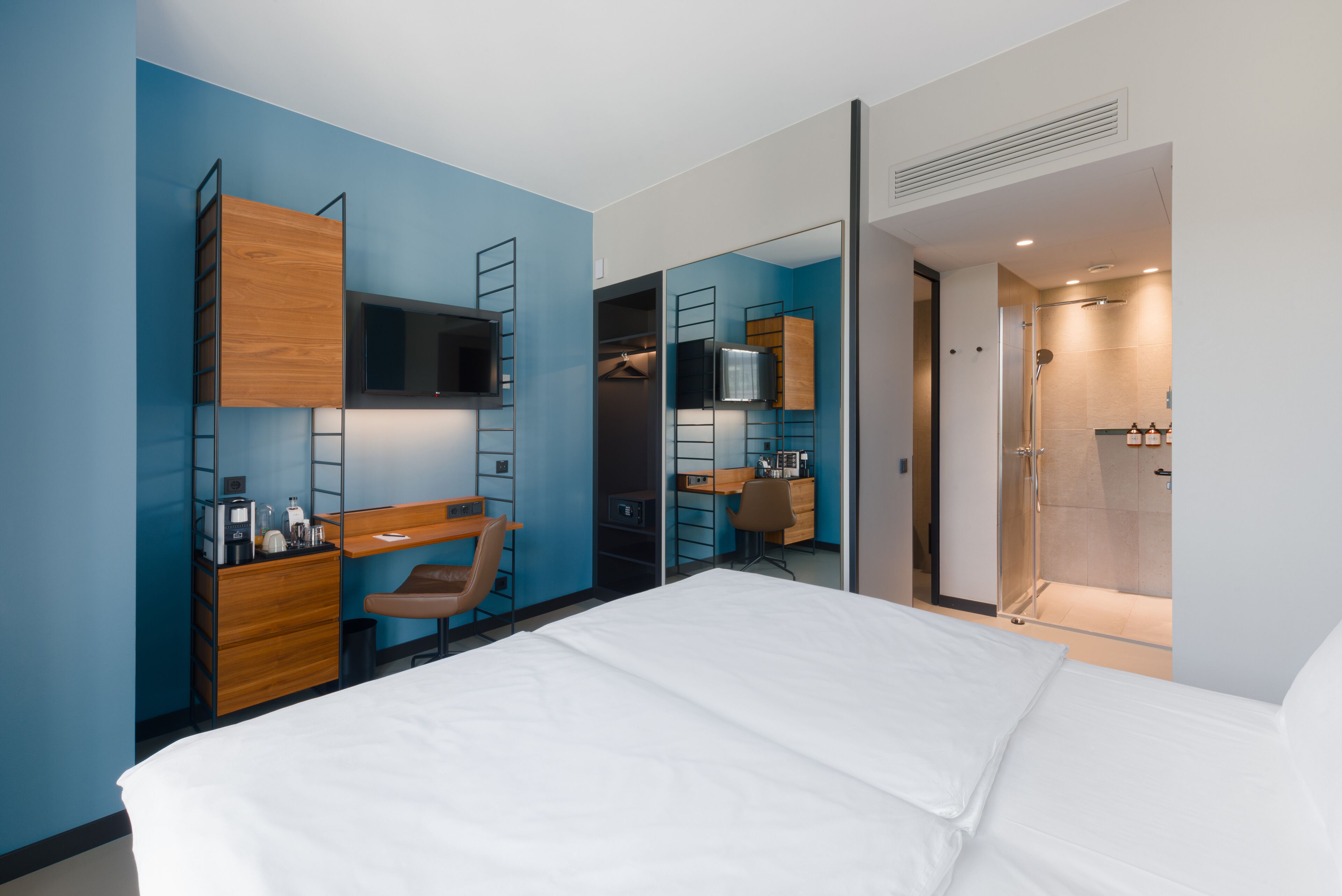 A modern hotel room with a bed, a desk, a chair, a wardrobe, and a shower area.
