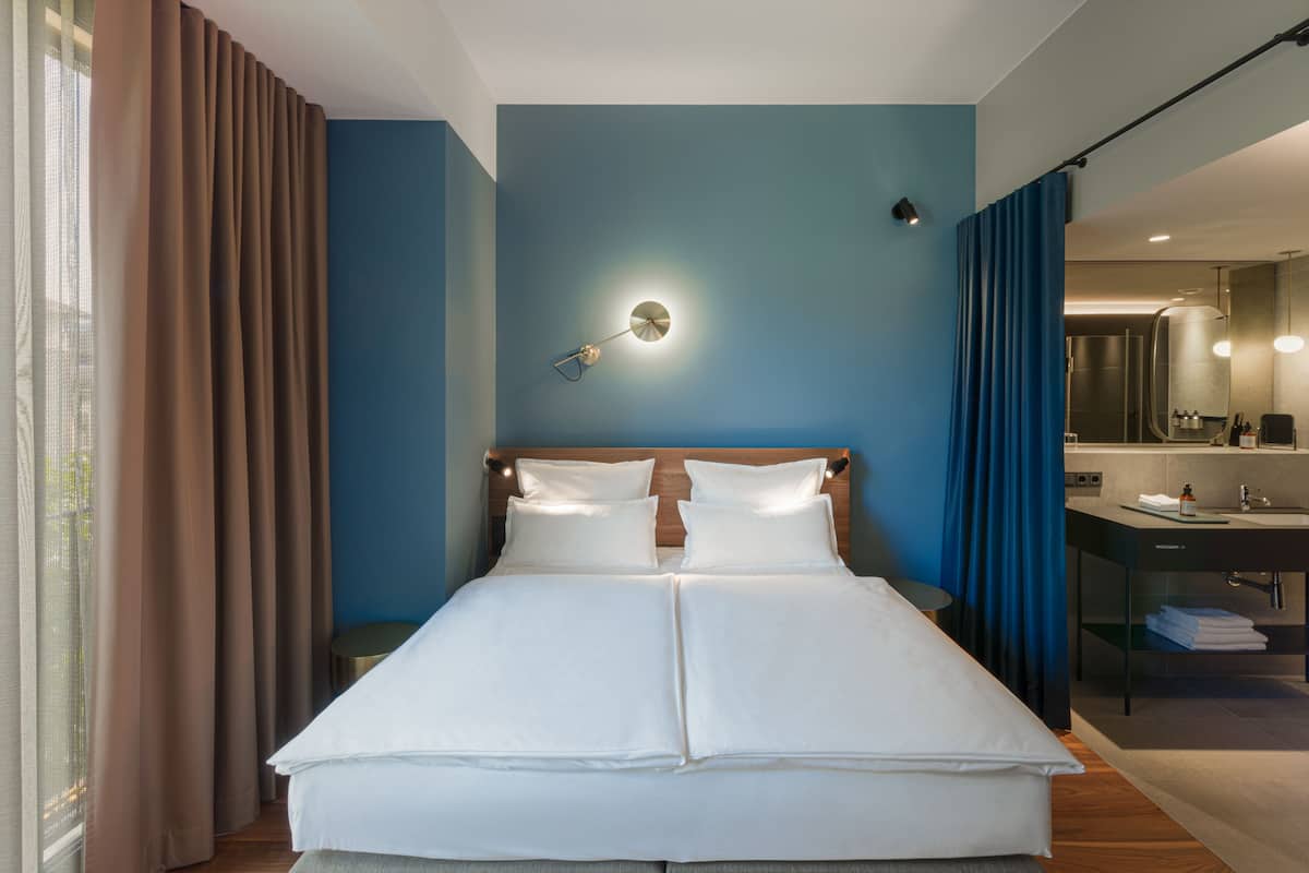 A neatly made bed in a room with a blue wall, a bedside table, and a bathroom visible through a glass partition.