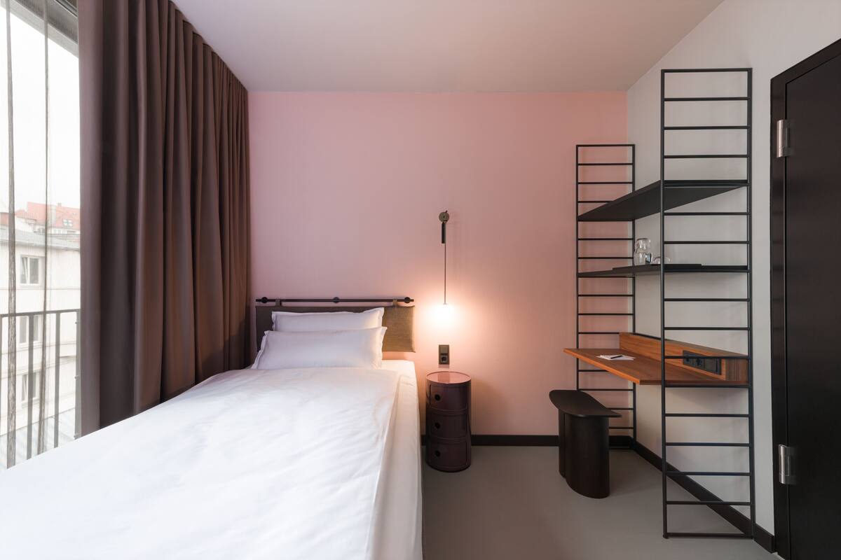 A hotel room with a bed, a desk with a chair, a small trash bin, and a wall-mounted shelf unit.