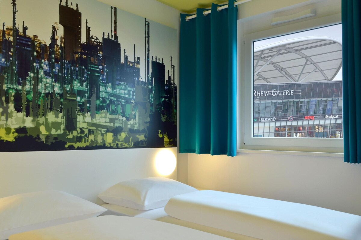 Habitación de hotel con cama, un cuadro de paisaje urbano, una ventana con cortinas y vistas a un edificio con letreros.