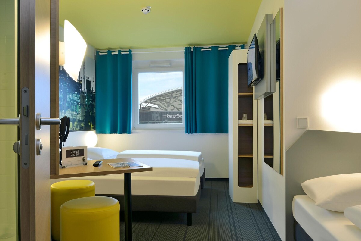 Una habitación de hotel compacta con una cama, un escritorio con silla, una mesita y un televisor montado en la pared.