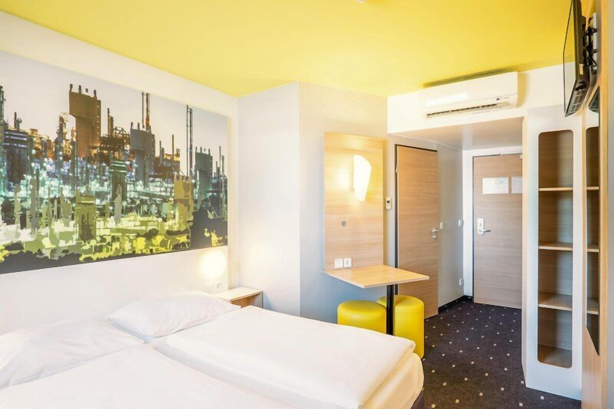Habitación de hotel con cama, escritorio, silla, armario y un mural con vista a la ciudad.
