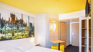 Habitación de hotel con una cama grande, un cuadro de un paisaje urbano, una mesita, una silla, un armario y un televisor.