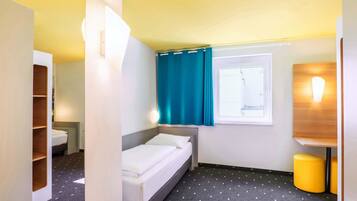 Habitación de hotel pequeña con cama, ventana con cortinas, mesita de noche, espejo y un taburete.