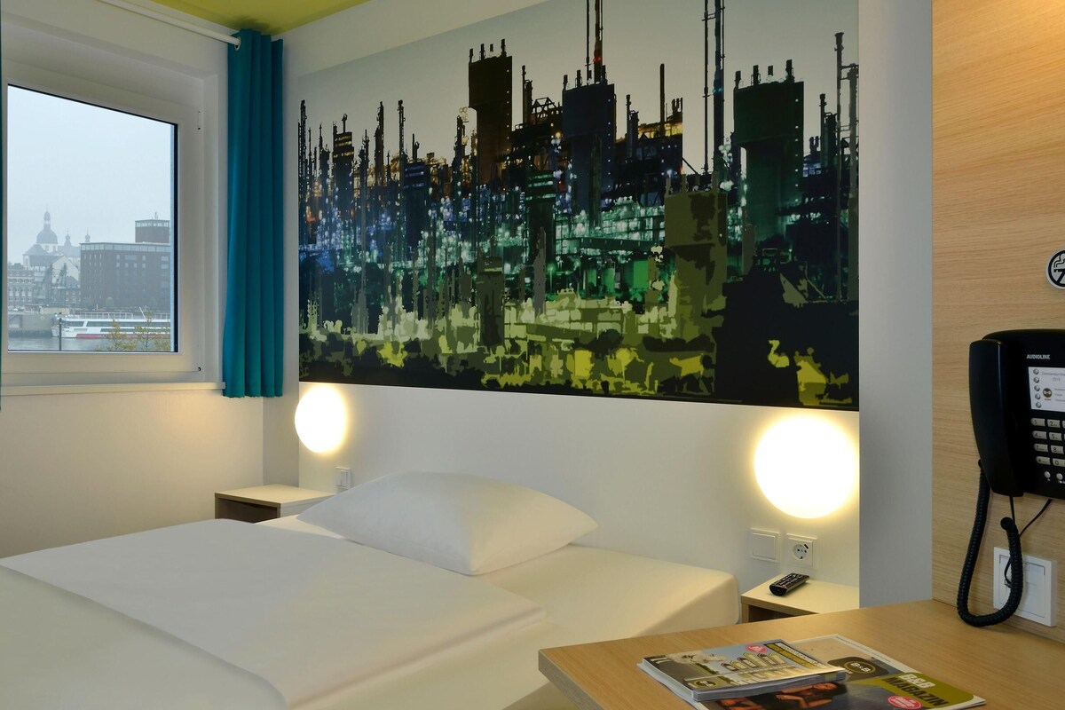 Habitación de hotel con cama, lámparas de noche, mesita de noche, teléfono en la pared y ventana con cortinas.