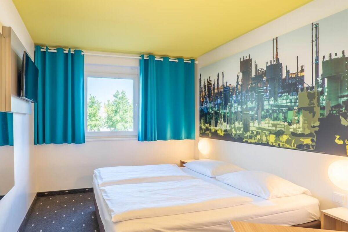 Habitación de hotel con dos camas, una ventana con cortinas, un cuadro de paisaje urbano y una pequeña mesa.