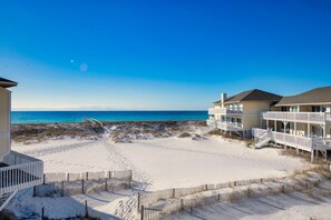 A beachfront property with a sandy beach, a wooden fence, and a building with a balcony overlooking the ocean. - Clean Beach front, On the sand Reno, FREE  Beach Service, Pools, Pickleball/Golf (Destin)
