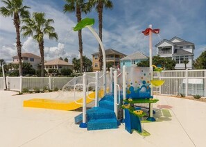 A water play area with slides and water jets, surrounded by palm trees and houses.