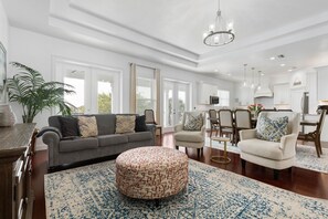 A spacious living room with a grey sofa, a round ottoman, and a dining area with a table and chairs.