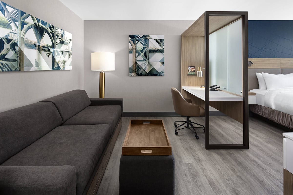 Une chambre d’hôtel moderne avec un canapé gris, une table d’appoint en bois, un bureau avec une chaise, un lit et des œuvres d’art abstraites accrochées aux murs.