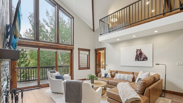 A spacious living area with a large window, a balcony, a high ceiling, and a cozy seating arrangement.